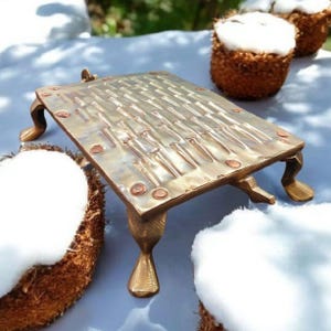 May include: A gold-colored, rectangular decorative tray with ornate legs and a textured surface. The tray is made of metal and has a vintage aesthetic. The tray is displayed with two halves of a coconut.