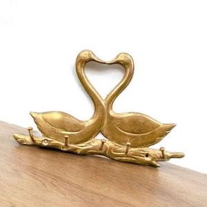 以下が含まれることがあります： 2 羽の白鳥がハート型を形成する、金色の金属製の壁掛けキーホルダー。 キーホルダーは、鍵を掛けるための 4 つのフックが付いた枝のようなベースを備えています。 白鳥は、質感のある羽で詳細に描かれています。