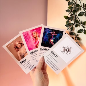 May include: Four album art prints are held in a hand. The prints feature images of female artists and album titles: "Amala", "Hot Pink", "Planet Her", and "Scarlet". Each print includes artist and release information.