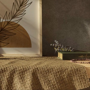 Op de afbeelding: Een ingelijst afdruk met een bruin en beige abstract ontwerp van een blad en cirkels. Een groen boek met een witte bloemtak erop ligt op een lichtbruin geweven deken. Een houten tafel met een bruine kralenketting bevindt zich op de voorgrond.