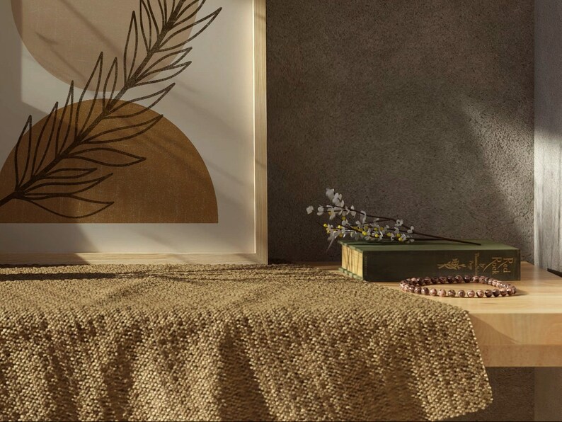 Op de afbeelding: Een ingelijst afdruk met een bruin en beige abstract ontwerp van een blad en cirkels. Een groen boek met een witte bloemtak erop ligt op een lichtbruin geweven deken. Een houten tafel met een bruine kralenketting bevindt zich op de voorgrond.