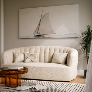 May include: A white, plush, curved sofa with a brown, glass coffee table in front of it. The sofa has several throw pillows in a variety of patterns. A painting of a sailboat on the water hangs on the wall behind the sofa.