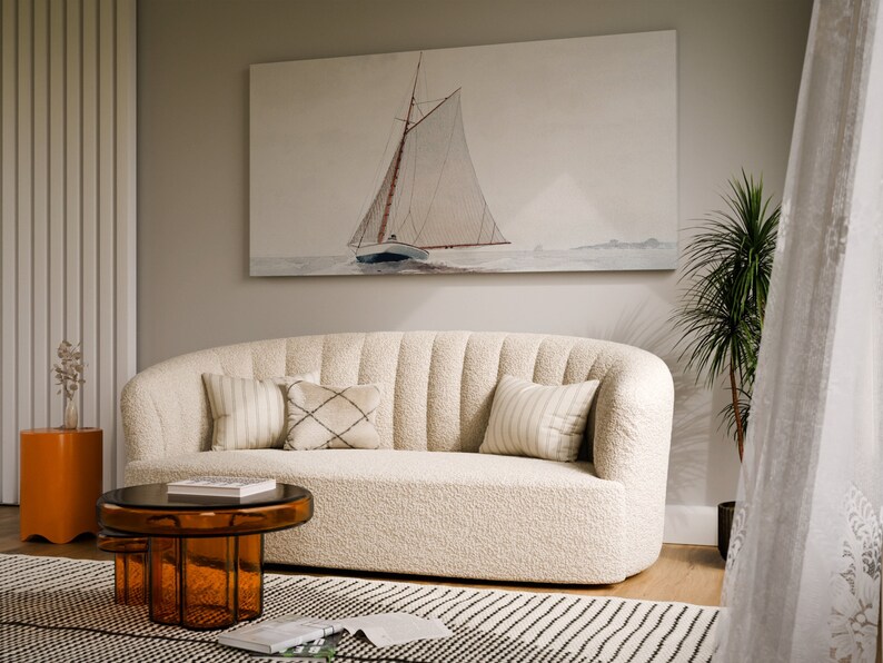 May include: A white, plush, curved sofa with a brown, glass coffee table in front of it. The sofa has several throw pillows in a variety of patterns. A painting of a sailboat on the water hangs on the wall behind the sofa.