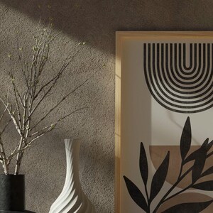 Op de afbeelding: Een ingelijst print met een zwart-wit abstract ontwerp met een reeks gebogen lijnen en een gestileerd blad. De print is op een cr&egrave;mekleurige achtergrond en is ingelijst in natuurlijk hout.