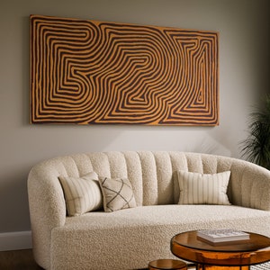 May include: A white, fluffy sofa with a curved back and three pillows. The sofa is in front of a wall with a large abstract painting featuring brown and orange lines. A small, round, orange side table is to the left of the sofa. A glass coffee table with a book on top is in front of the sofa.