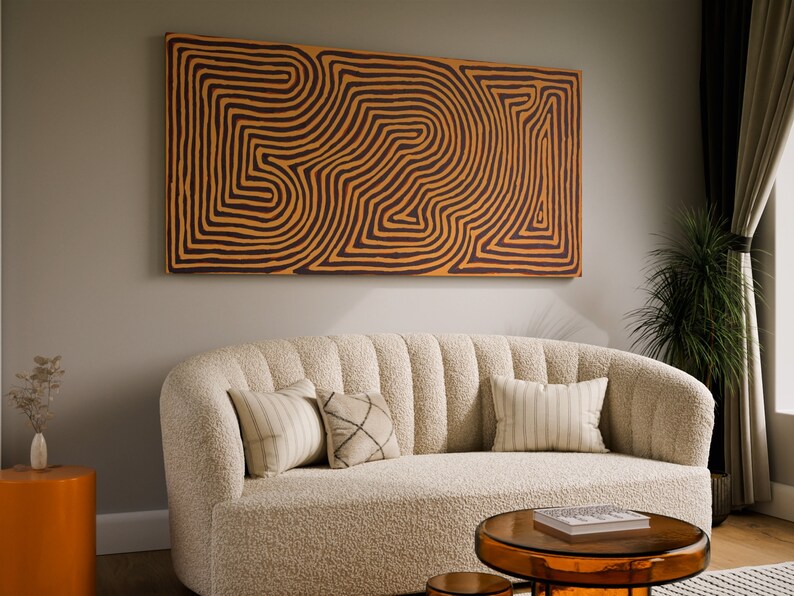 May include: A white, fluffy sofa with a curved back and three pillows. The sofa is in front of a wall with a large abstract painting featuring brown and orange lines. A small, round, orange side table is to the left of the sofa. A glass coffee table with a book on top is in front of the sofa.