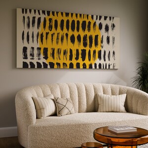 May include: A white, fluffy sofa with a black and yellow abstract painting hanging above it. The sofa has three pillows, one with a striped pattern, one with a geometric pattern, and one with a solid color. There is a small, round, orange side table to the left of the sofa and a glass coffee table in front of the sofa.