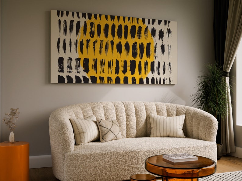May include: A white, fluffy sofa with a black and yellow abstract painting hanging above it. The sofa has three pillows, one with a striped pattern, one with a geometric pattern, and one with a solid color. There is a small, round, orange side table to the left of the sofa and a glass coffee table in front of the sofa.