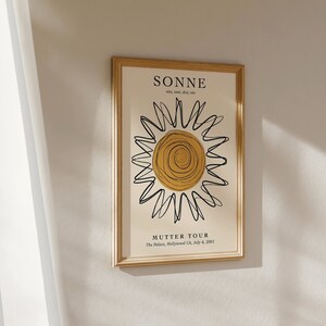 May include: A framed print with a gold frame and a white background. The print features a black and yellow sun with the text "SONNE" and "MUTTER TOUR" in black.