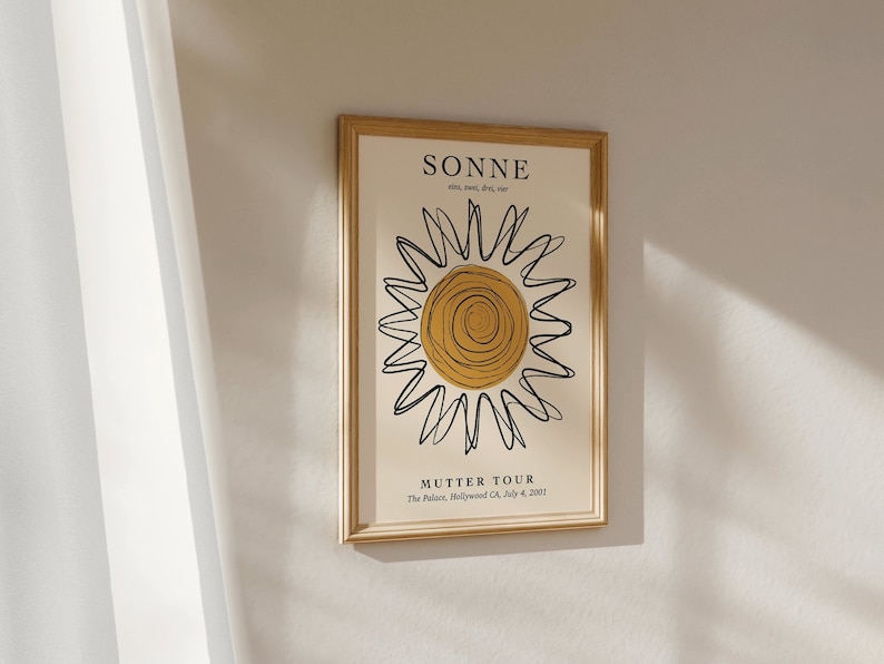 May include: A framed print with a gold frame and a white background. The print features a black and yellow sun with the text "SONNE" and "MUTTER TOUR" in black.