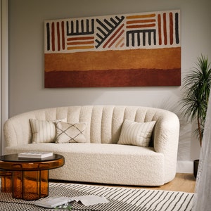May include: A white, fluffy, curved sofa with pillows sits in a living room with a brown and orange abstract painting on the wall, a brown coffee table, and a small orange side table.