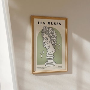May include: A framed print with a gold frame and a white background. The print features a line drawing of a bust of a person with the text "LES MUSES" at the top. The text "EXPOSITION TEMPORELLE" and "GALERIE LIMITAIRE" are at the bottom left, and "PARIS 2023" and "COLLECTION LES MUSES" are at the bottom right.
