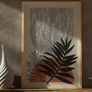 Op de afbeelding: Een ingelijst print met een minimalistisch ontwerp met zwarte en witte abstracte lijnen en een gestileerd zwart blad op een bruine achtergrond.