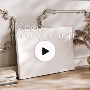 May include: A white canvas with the text "Video mockup" and a play button icon. The canvas is propped against a wall with decorative moulding. Dried plants and a stack of books are also present.