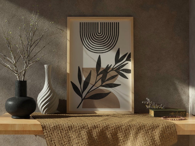 Op de afbeelding: Een ingelijste print met een geometrisch ontwerp in zwart en wit met een boog en een gestileerd blad. De print is op een witte achtergrond en is ingelijst in een naturel houten lijst. De print staat op een houten tafel met een bruin geweven kleed ervoor.