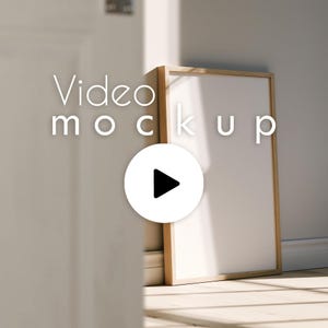以下が含まれることがあります： 白い内側の長方形の木製フォトフレームが壁に寄りかかっています。「Video mockup」というテキストが白で表示され、画像には再生ボタンのアイコンが重ねられています。フレームは日当たりの良い部屋に配置されています。