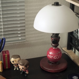 Puede incluir: Una pantalla de lámpara blanca con una base roja con temática de Coca-Cola. La lámpara está sobre una superficie negra con dos pequeñas figuras.
