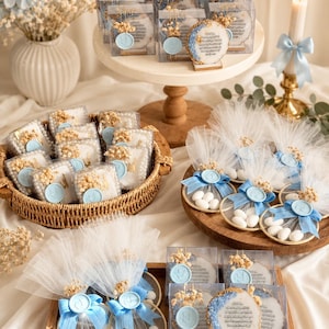 May include: A collection of party favors featuring round resin plaques with gold edges and Arabic calligraphy, clear square boxes, and circular gold frames with white sugared almonds, all accented with light blue wax seals and dried flowers.