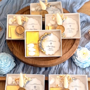 Puede incluir: Un conjunto de cinco cajas de regalo blancas con tapas transparentes, cada una contiene un pequeño disco de cerámica blanca con detalles dorados, una pequeña caja blanca con tres almendras blancas, una pequeña caja blanca con un sello de cera dorado y un pequeño ramo de flores secas. Los discos de cerámica tienen el texto "Ariel Urena Bautizo 03 Agosto 2025" impreso en ellos.