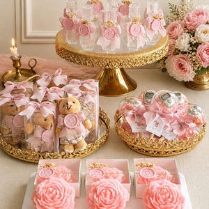 May include: A collection of party favors including clear glass bottles with pink ribbons and wax seals, plush teddy bears in clear boxes, clear spheres with digital counters, and pink rose-shaped candles on a white marble tray with gold accents.