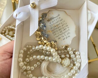 May include: A white gift box with a white ribbon and bow. Inside the box is a white prayer bead necklace with a white tassel and a white and gold decorative piece with Arabic text. The text reads: "Allah la ilaha illa huwa al-hay al-qayyum la ta'khuzuhu sinatu wa la nawm lahu ma fi al-samawati wa al-ard man dhalika alladhi yashfa'u 'indahi illa bi-idhnhi ya'lamu ma bayna aydihim wa ma khalfahum wa la yuhithuna bi-shay'in min 'ilmihi illa bi-ma sha'a wasi'a kursiyyuhu al-samawati wa al-ard wa la ya'uduhu hifzhuhuma wa huwa al-'aliyyu al-'azim"