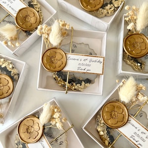 May include: Small white boxes with clear lids containing gold and black resin coasters with dried flowers and a wax seal. The coasters have a handwritten message that says "Luciana's 40th Birthday 10/20/2022 Thank you".
