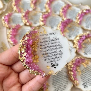 May include: A white, heart-shaped resin ornament with gold and pink accents. The ornament features an Arabic inscription and the word "Sıla" in black lettering.