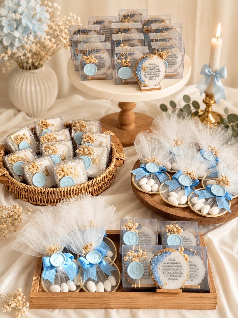 May include: A collection of party favors featuring round resin plaques with gold edges and Arabic calligraphy, clear square boxes, and circular gold frames with white sugared almonds, all accented with light blue wax seals and dried flowers.
