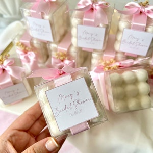 Op de afbeelding: Roze en witte cadeauboxen gevuld met witte marshmallows, gebonden met roze lint en versierd met kleine witte bloemetjes. De dozen hebben een wit label met de tekst "Mary's Bridal Shower 06.07.24".