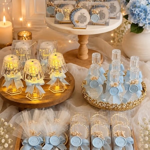 Op de afbeelding: Een verzameling feestartikelen met doorzichtige, lampvormige acryllampen met de naam Faisal, glazen parfumflesjes met blauwe waszegels en ronde acrylplaten met gedroogde bloemen, allemaal geaccentueerd met lichtblauwe linten.