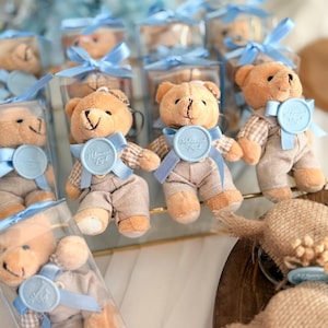 Puede incluir: Pequeños osos de peluche en cajas transparentes, atados con cintas azul claro. Los osos visten trajes color beige y tienen medallones azules. El fondo incluye más osos en caja y un arreglo floral borroso.