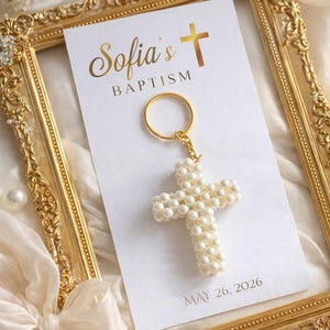 May include: A pearl cross keyring with a gold ring and chain, on a white card with "Sofia's Baptism" and "May 26, 2026". The card is in a gold ornate frame. The image has a soft, elegant aesthetic.