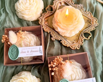 May include: Two white rose-shaped candles in clear boxes with brown lids. The boxes are decorated with dried flowers and a green wax seal with a rose design. The text on the boxes reads "Thank you for celebrating Jesse & Jovonia 08.23.2025".