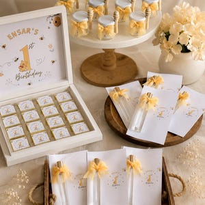 Pode incluir: Uma caixa de madeira branca contendo dezasseis chocolates quadrados com etiquetas personalizadas, pequenos frascos de mel de vidro com colheres de pau e laços amarelos num pedestal, e frascos de vidro com laços amarelos num tabuleiro de madeira.