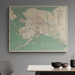 May include: A vintage map of Alaska with a green background and red and black text. The map shows the state's major cities, rivers, and mountains. The map is framed and hanging on a grey wall.