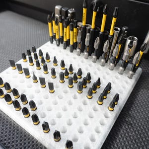 May include: A white foam organizer filled with various black and yellow screwdriver bits and attachments. The tools are arranged in a grid pattern, with some standing upright and others lying flat. The tools are for construction and repair.