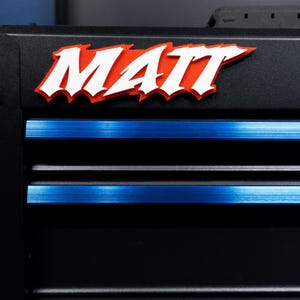 May include: Close-up of a black toolbox with blue accents. The word "MATT" is displayed in white, outlined in red, and has a stylized design. The toolbox has horizontal blue stripes and a black background.