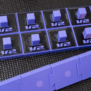 May include: A set of black and purple square modular components featuring a raised purple cube in the centre and the text 1/2 printed on the front. Below these are interlocking purple rectangular pieces with circular indentations on each.