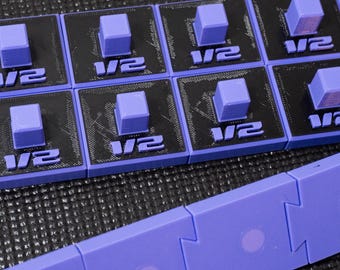 May include: A set of black and purple square modular components featuring a raised purple cube in the center and the text 1/2 printed on the front. Below these are interlocking purple rectangular pieces with circular indentations on each.