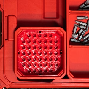 May include: A bright red plastic tool storage case featuring a square insert with a grid of hexagonal holes for organising bits. To the right, a separate compartment contains a collection of loose silver metal screwdriver bits of various sizes.