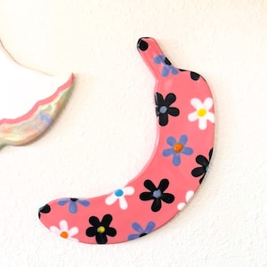 May include: A pink banana-shaped decorative item adorned with a pattern of black, white, and blue flowers. The banana has a glossy finish and is set against a white textured background. The design is whimsical and playful.