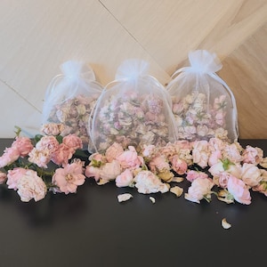 May include: Three white organza bags filled with dried pink rose petals. The bags are sitting on a wooden surface. There are also loose dried pink rose petals scattered on a black surface.