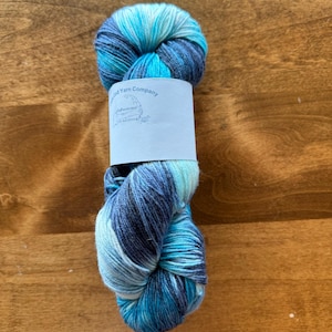 Könnte beinhalten: Eine Knäuel blauer, weißer und grauer Wolle mit einem Etikett, auf dem "The Wooded Yarn Company" steht.