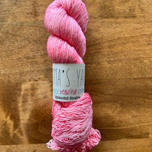 Op de afbeelding: Een bol roze en wit garen met een label waarop staat "Emma's Yarns Crazy Beautiful Colors Splendid Singles".