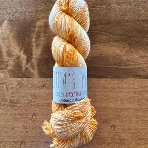 Op de afbeelding: Een bol oranje en witte garen met een label met de tekst "Emma's Yarn, Crazy Beautiful Color, Splendid Singles".