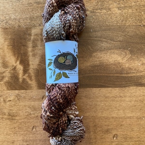 May include: A skein of brown and gray yarn with brown beads, wrapped around a white label with a bird's nest illustration and the text "MADE BY BIRD" and "hand dyed in the USA".