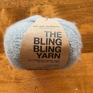 May include: A ball of light blue yarn labeled "The Bling Bling Yarn" with the text "we are knitters" and "weareknitters.com". The yarn is 79% superwash merino wool, 18% mohair, and 3% lurex. It weighs 1.75 ounces and is 230 yards long.