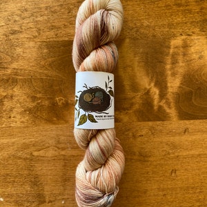 May include: A skein of hand-dyed yarn in shades of brown, cream, and beige. The yarn is wrapped around a white label with a bird's nest illustration and the text "MADE BY HEART & SOUL hand dyed in the Pacific Northwest".