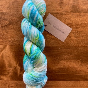 May include: A vibrant skein of yarn, featuring a mix of blue, white, and green shades. The yarn is twisted and has a tag that reads "SOCK OBSESSION YARNS". It's presented on a wooden surface.