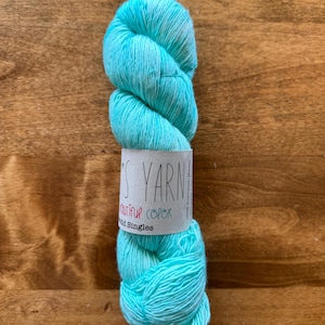 Op de afbeelding: Een bol lichtblauw garen met een wit label waarop "The's Yarn Beautiful Color Hand Singles" staat.